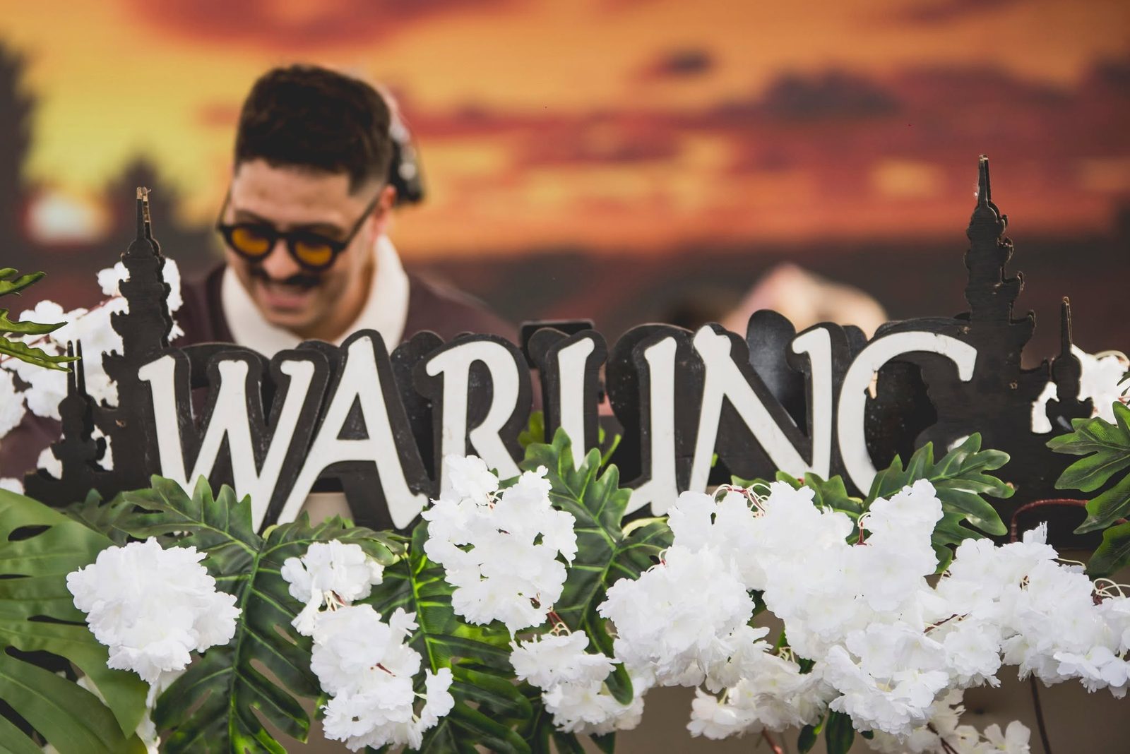 Kevin Disko sorrindo atrás do letreiro WARUNG no pôr do sol